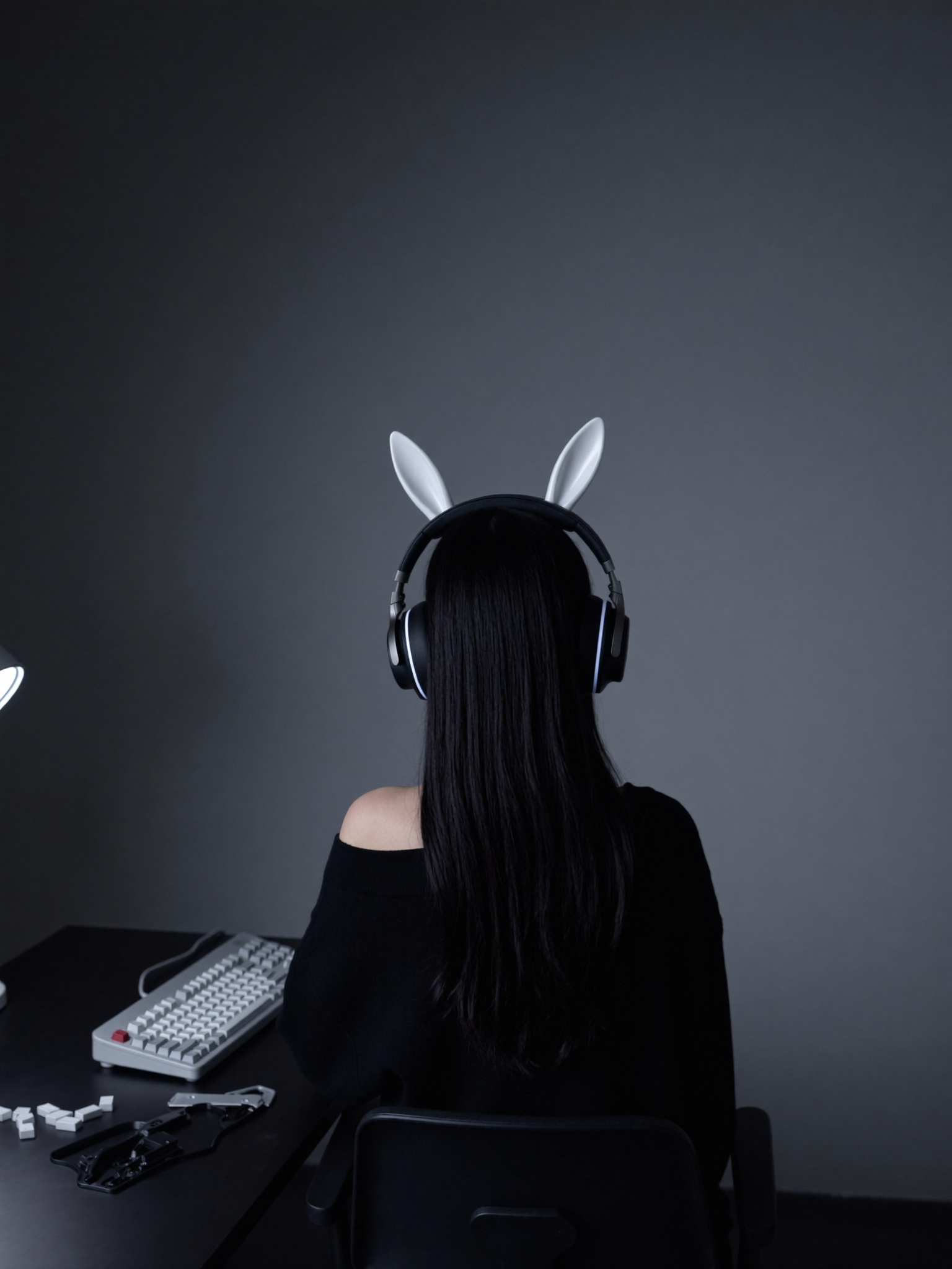 A young woman sits alone in a dark, nearly empty room, her back to the camera, in front of a keyboard she has taken apart.