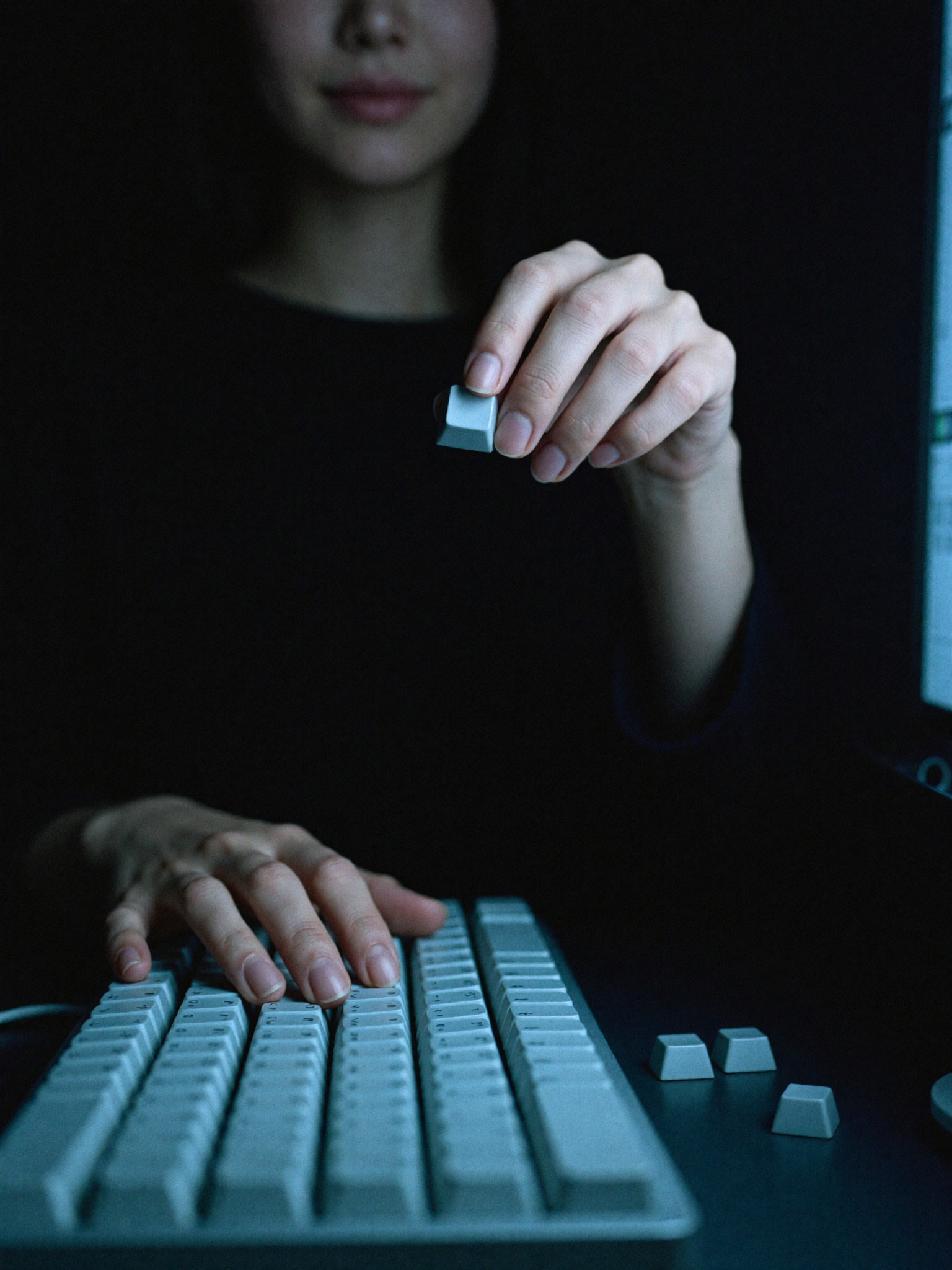 Close view of two hands over a mechanical keyboard in near darkness, one hand lifting a single keycap off the board.